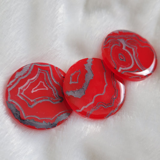 Three bright red slightly translucent round epoxy resin pieces with engraved details that resemble geode layers in shades of silver and grey.