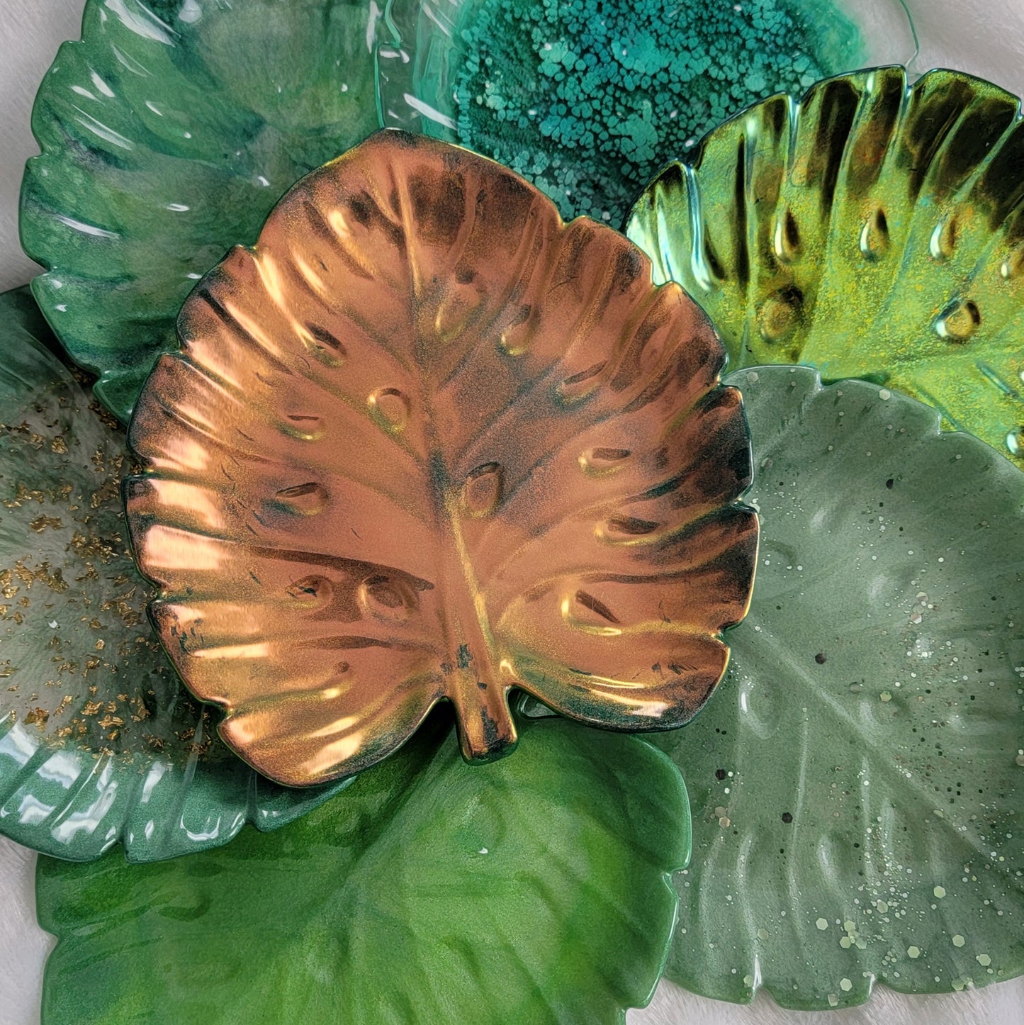 Variety of monstera leaf shaped decorative trays, shown in classic grass green, iridescent gold , gold foil flake and dark green, light sage and glitter, marbled green, chameleon green, and green ink.