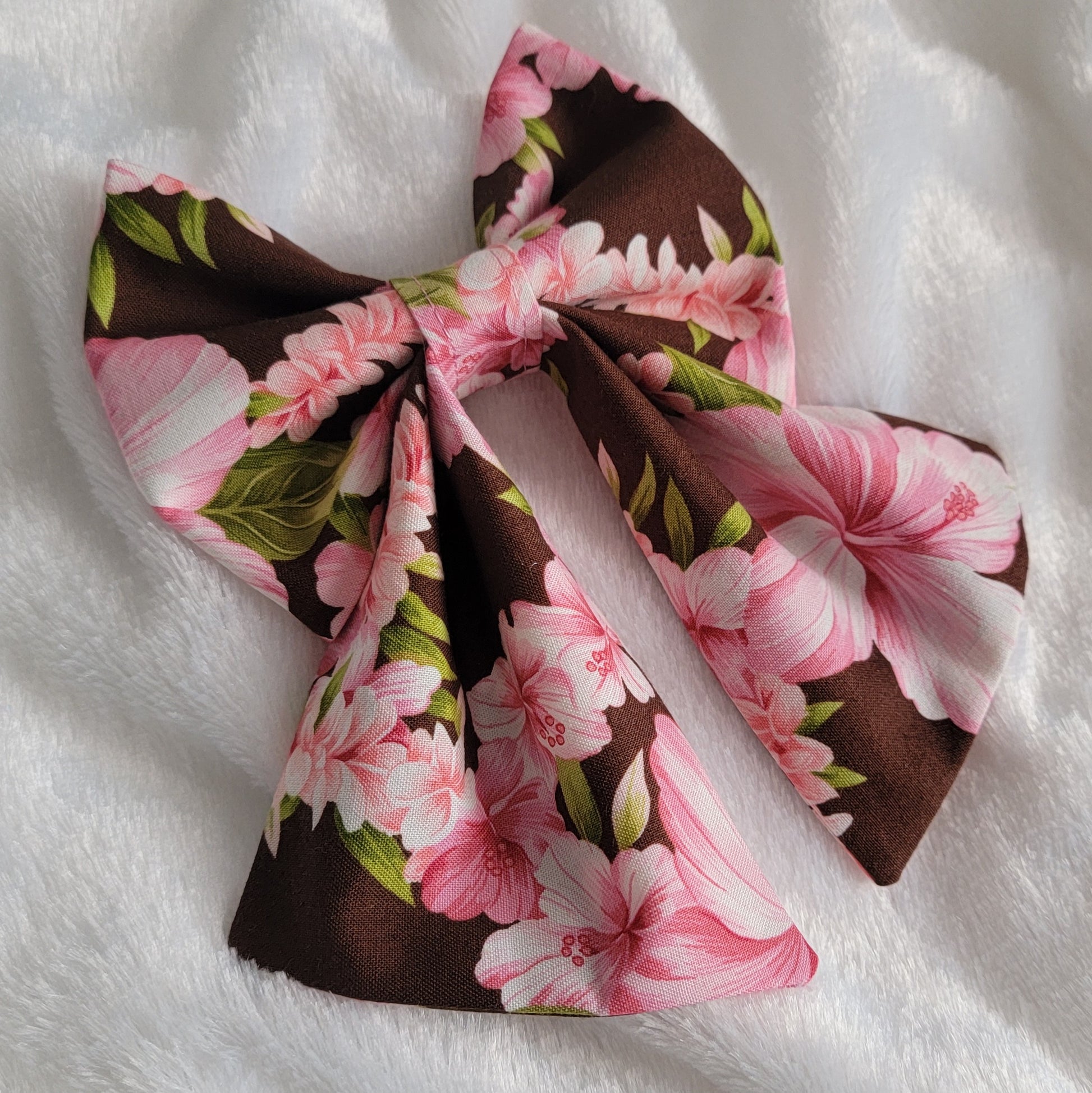 A fabric pet bow with a floral pattern of pink hibiscus leis on rich brown background, sailor bow style with long showy tails.