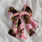 A fabric pet bow with a floral pattern of pink hibiscus leis on rich brown background, sailor bow style with long showy tails.
