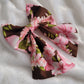 A fabric pet bow with a floral pattern of pink hibiscus leis on rich brown background, sailor bow style with long showy tails.