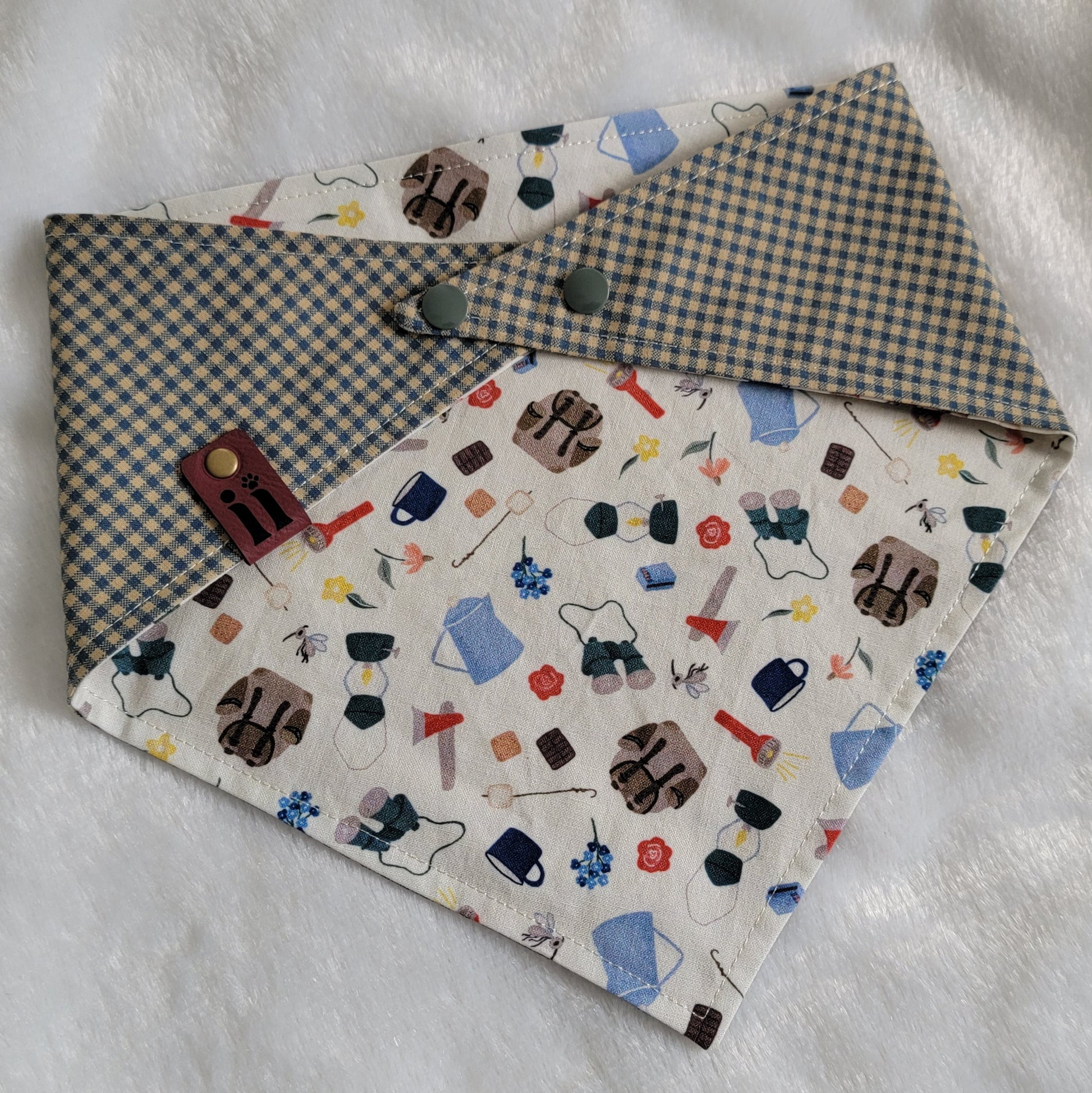 A reversible pet bandana with one side featuring an assortment of camping-themed patterns including a tent, campfire, and backpack, and the other side in a blue gingham plaid. 