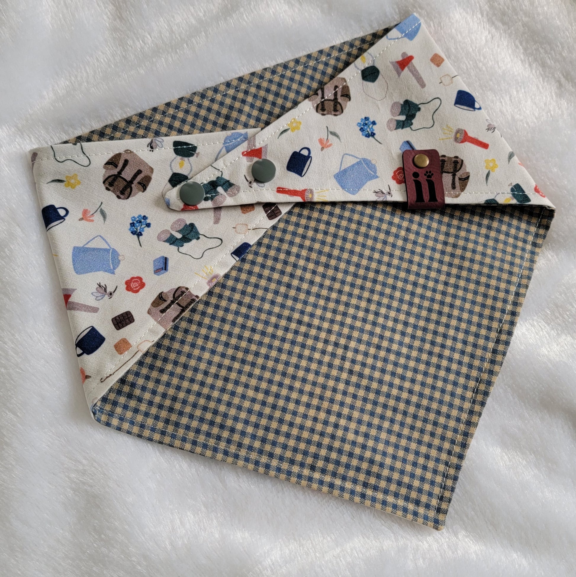 A reversible pet bandana with one side featuring an assortment of camping-themed patterns including a tent, campfire, and backpack, and the other side in a blue gingham plaid. 
