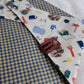 A reversible pet bandana with one side featuring an assortment of camping-themed patterns including a tent, campfire, and backpack, and the other side in a blue gingham plaid. 