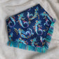 A blue bandana with a mermaid pattern and vibrant blue and rainbow fringe edges.