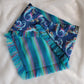 A blue bandana with a mermaid pattern and vibrant blue and rainbow fringe edges.