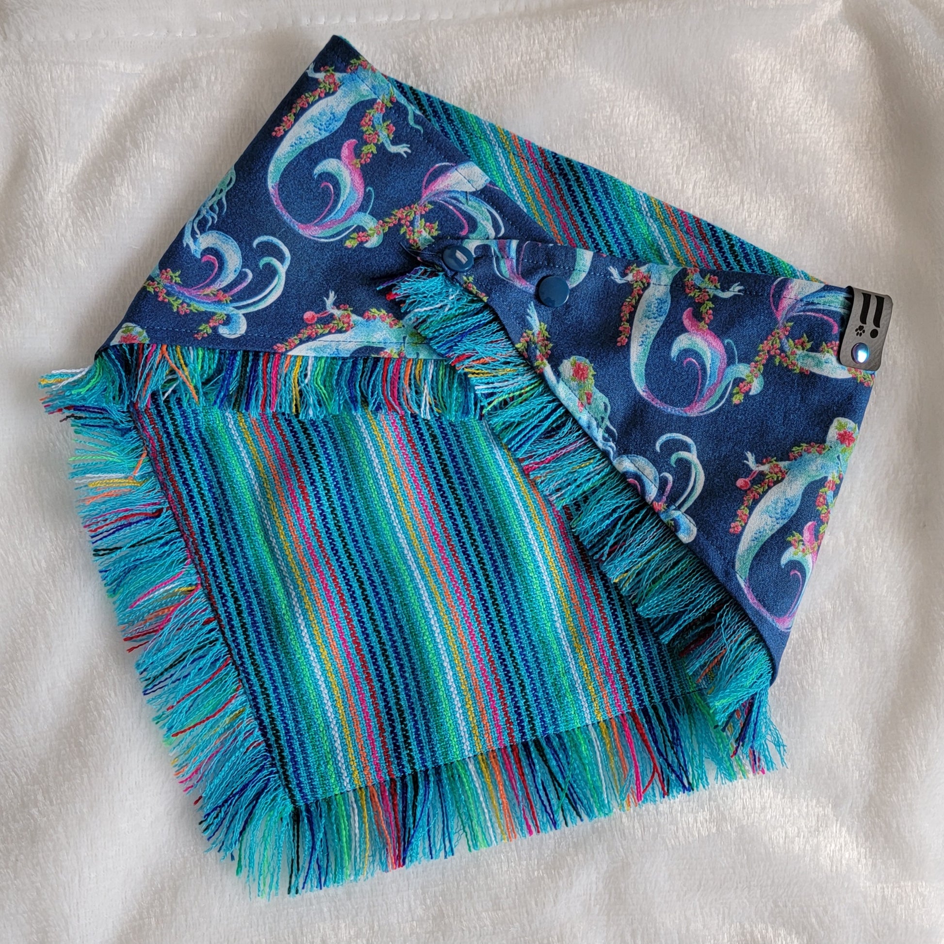 A blue bandana with a mermaid pattern and vibrant blue and rainbow fringe edges.