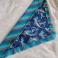 A blue bandana with a mermaid pattern and vibrant blue and rainbow fringe edges.