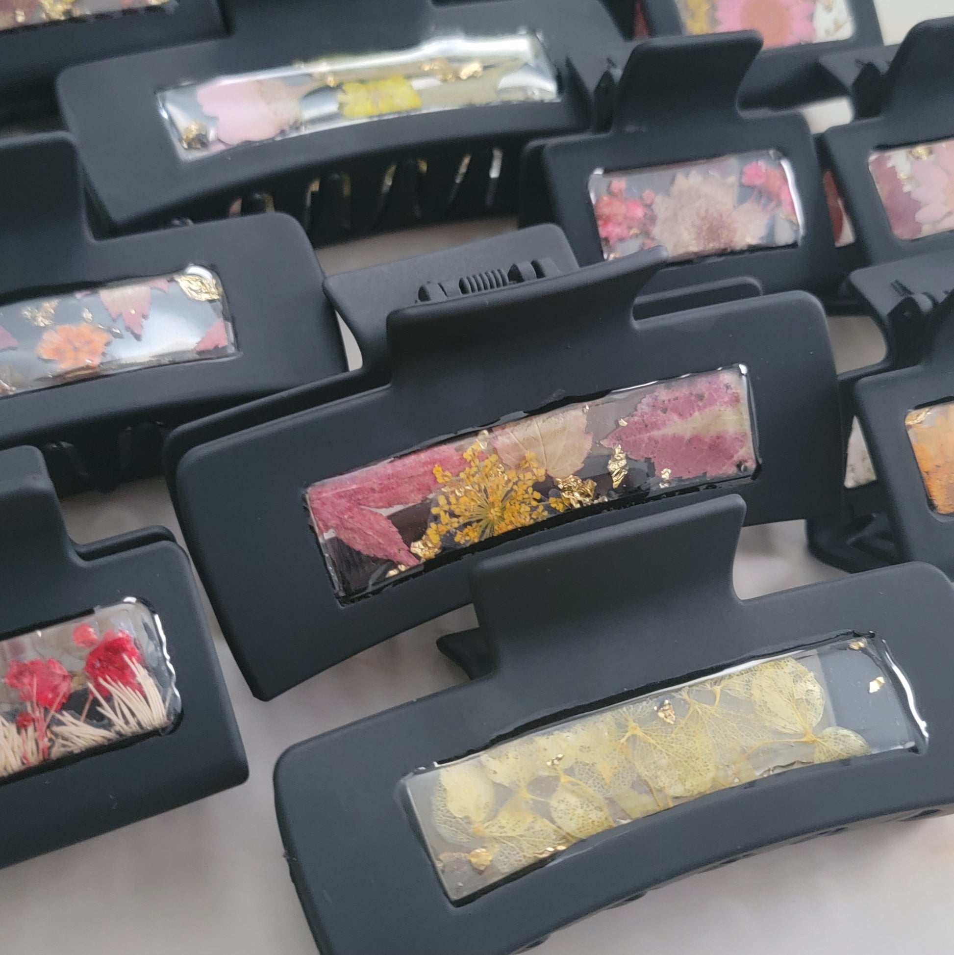 An array of 4-inch and 2-inch black claw clips with open spaces that are filled with pressed flowers and foliage and gold flakes cast in resin.