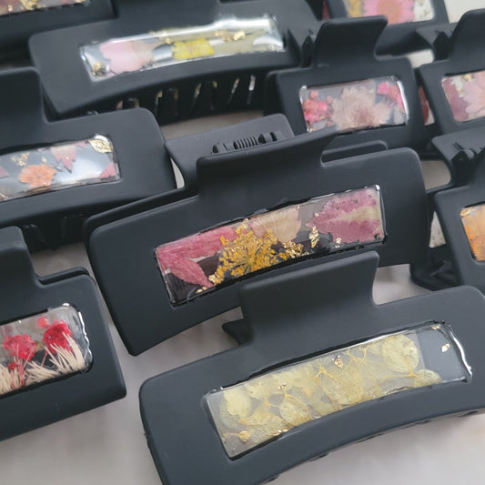 An array of 4-inch and 2-inch black claw clips with open spaces that are filled with pressed flowers and foliage and gold flakes cast in resin.