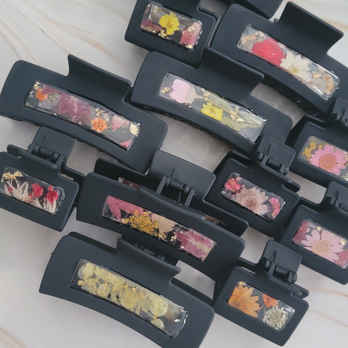 An array of 4-inch and 2-inch black claw clips with open spaces that are filled with pressed flowers and foliage and gold flakes cast in resin.