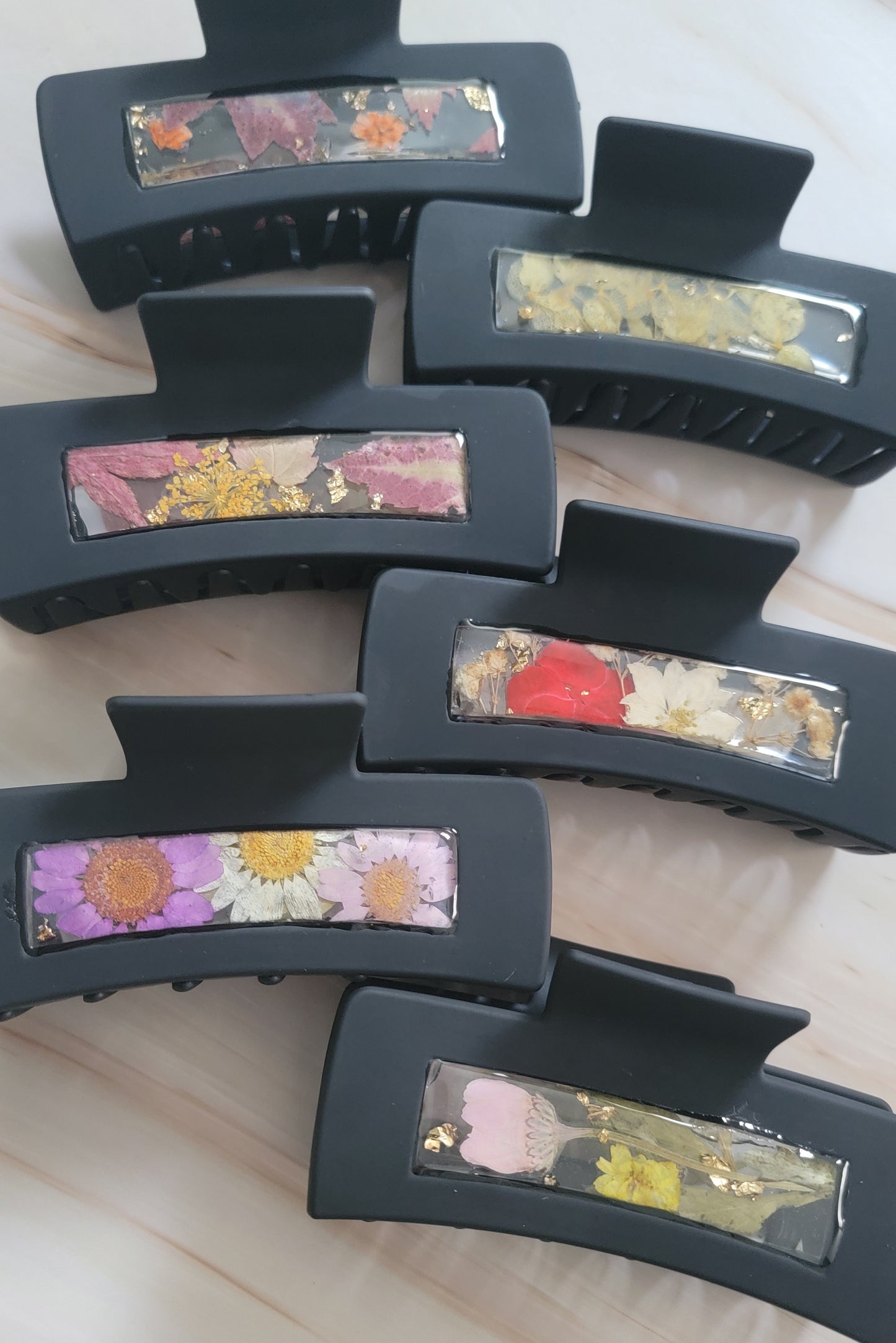 Six black 2-inch claw clips with open spaces that are filled with pressed flowers and foliage and gold flakes cast in resin.