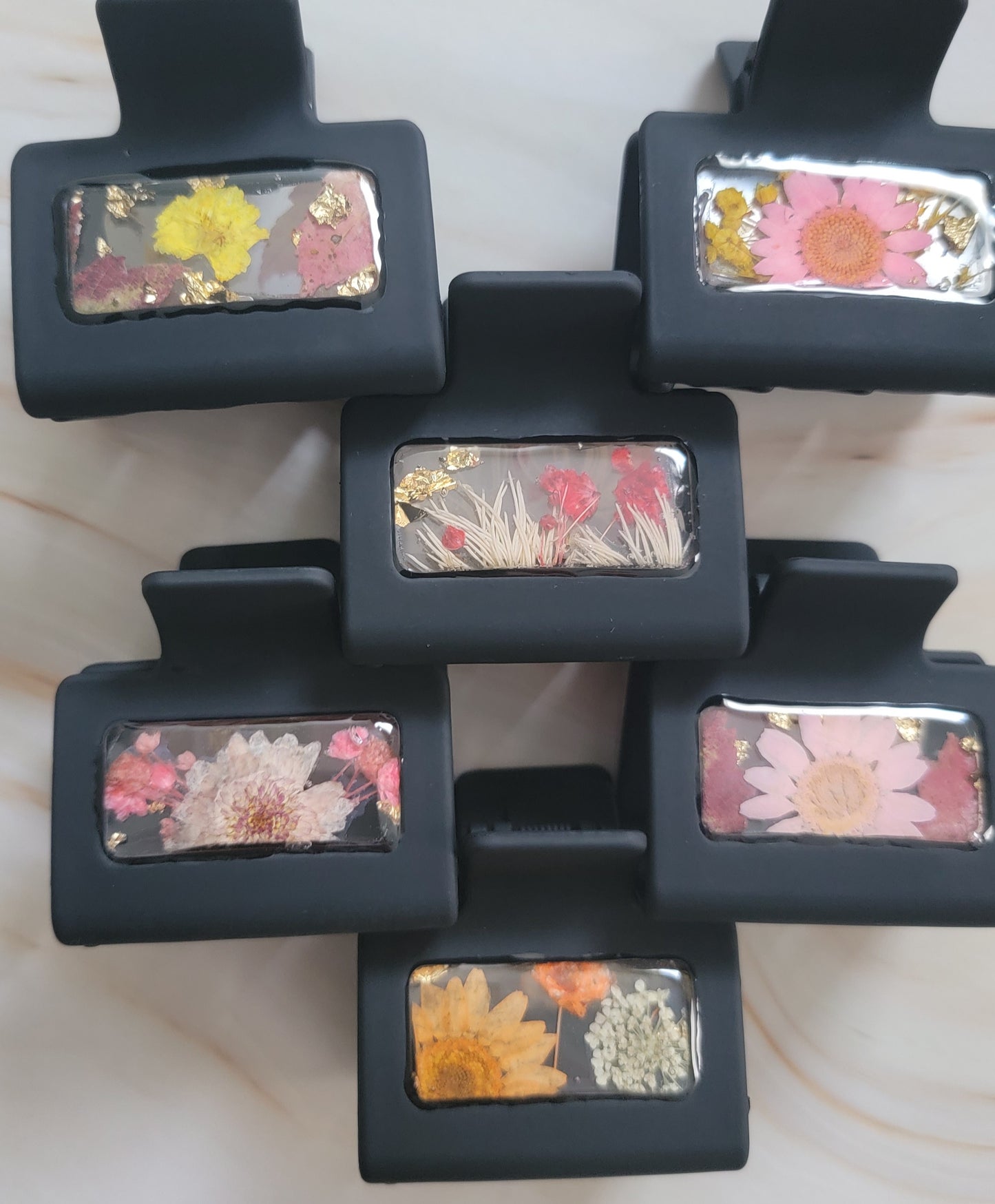 Six black 2-inch claw clips with open spaces that are filled with pressed flowers and foliage and gold flakes cast in resin.