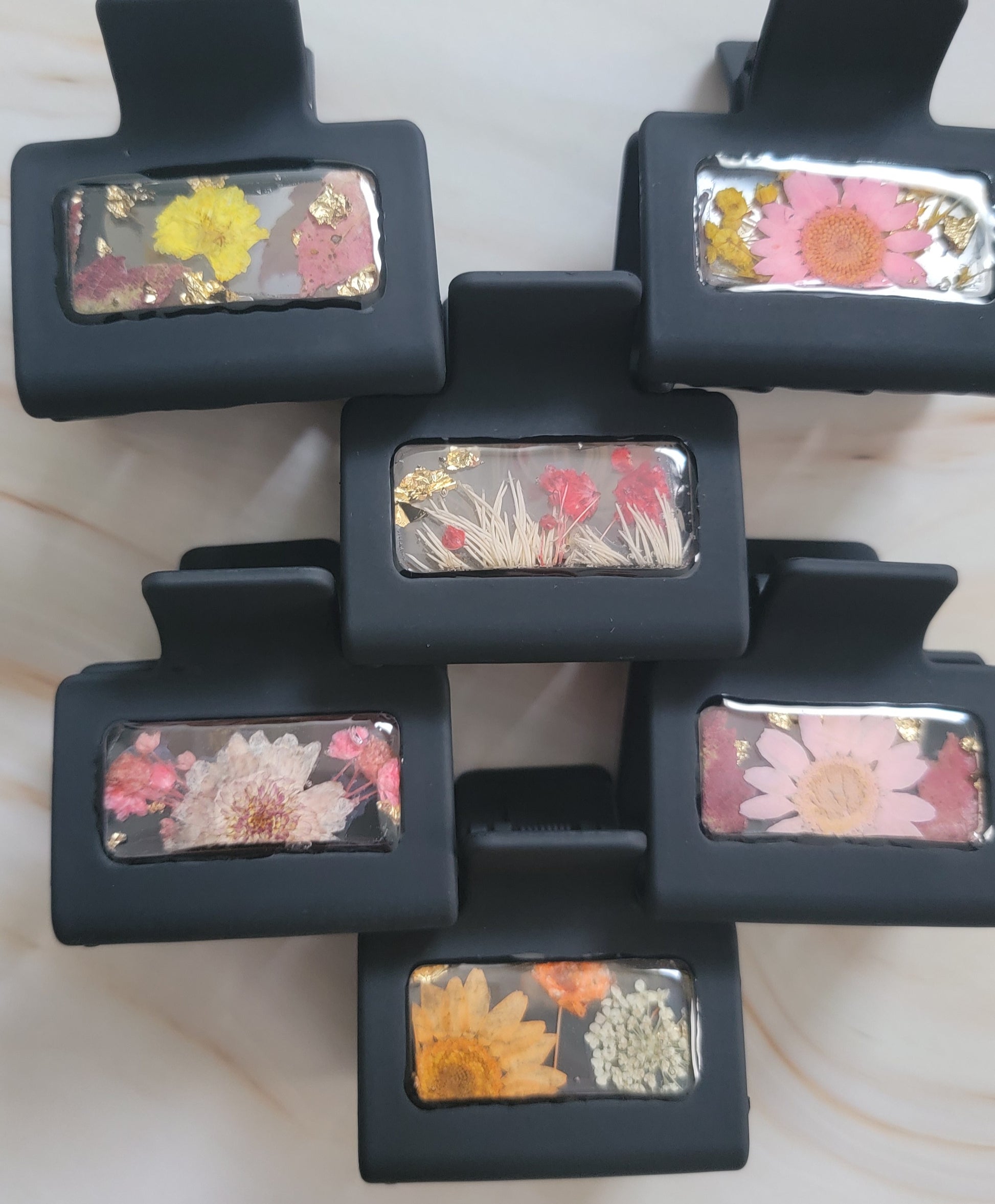 Six black 2-inch claw clips with open spaces that are filled with pressed flowers and foliage and gold flakes cast in resin.
