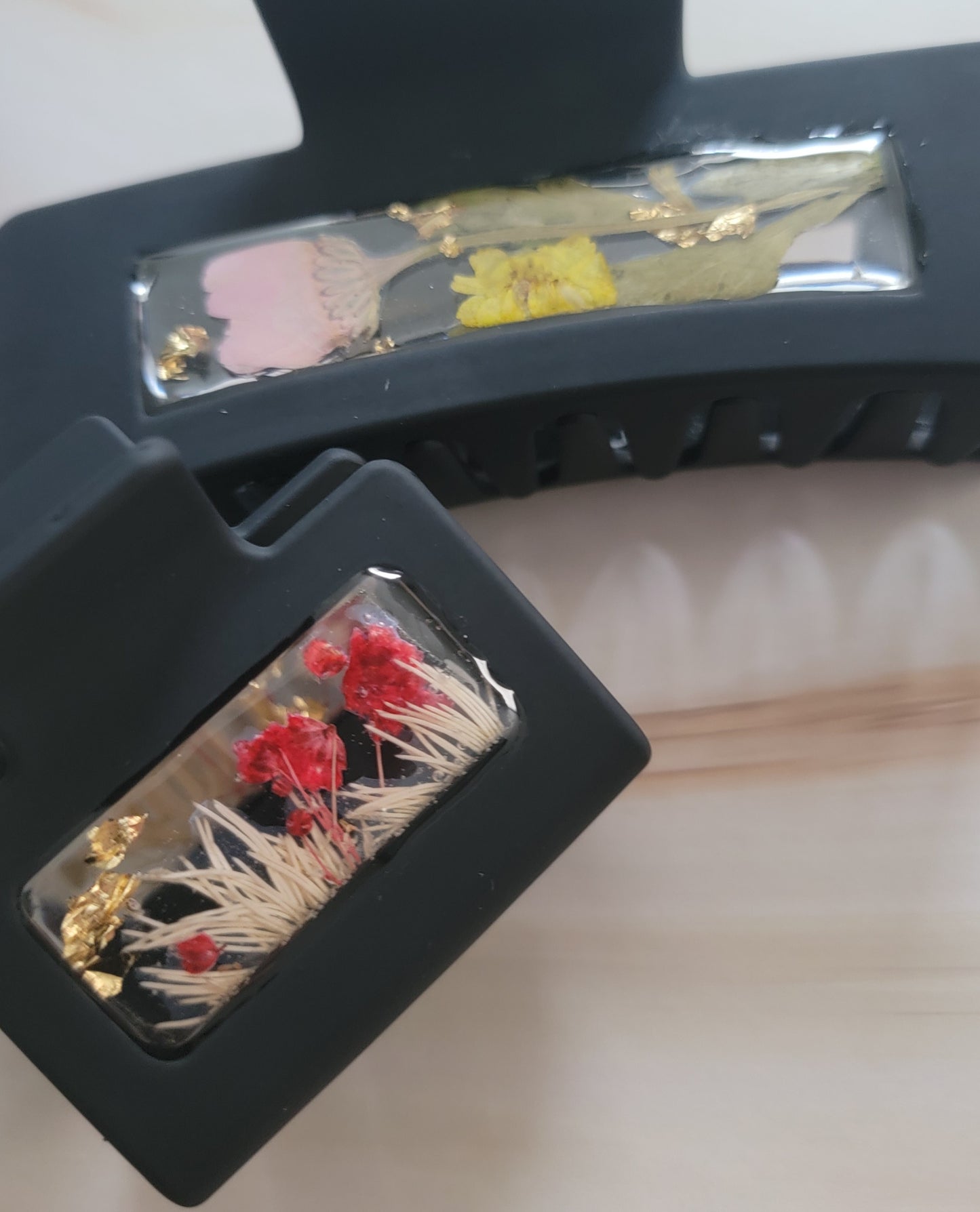 An array of 4-inch and 2-inch black claw clips with open spaces that are filled with pressed flowers and foliage and gold flakes cast in resin.