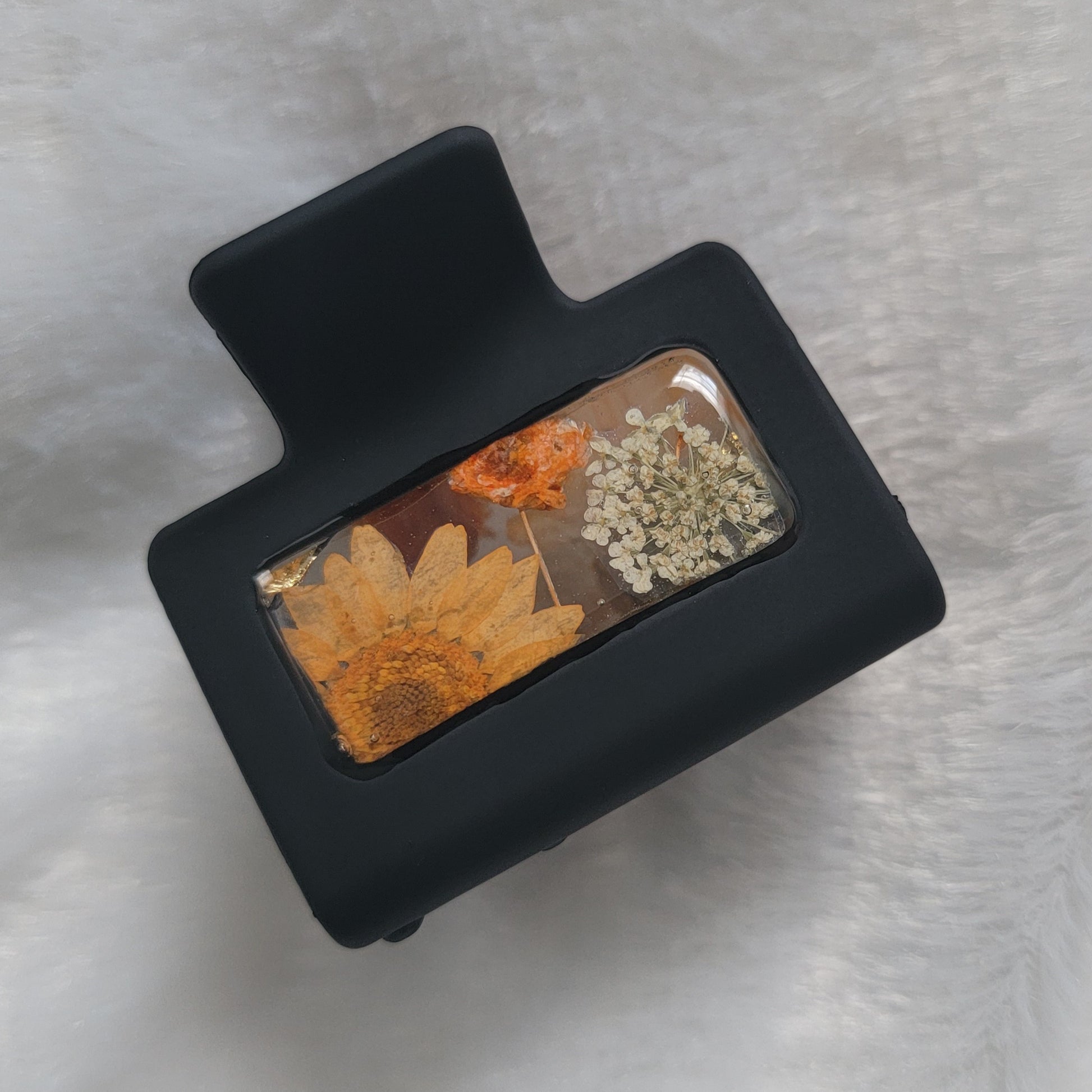 A black 2-inch claw clip with orange and white pressed flowers cast in resin within an open space in the hair clip.