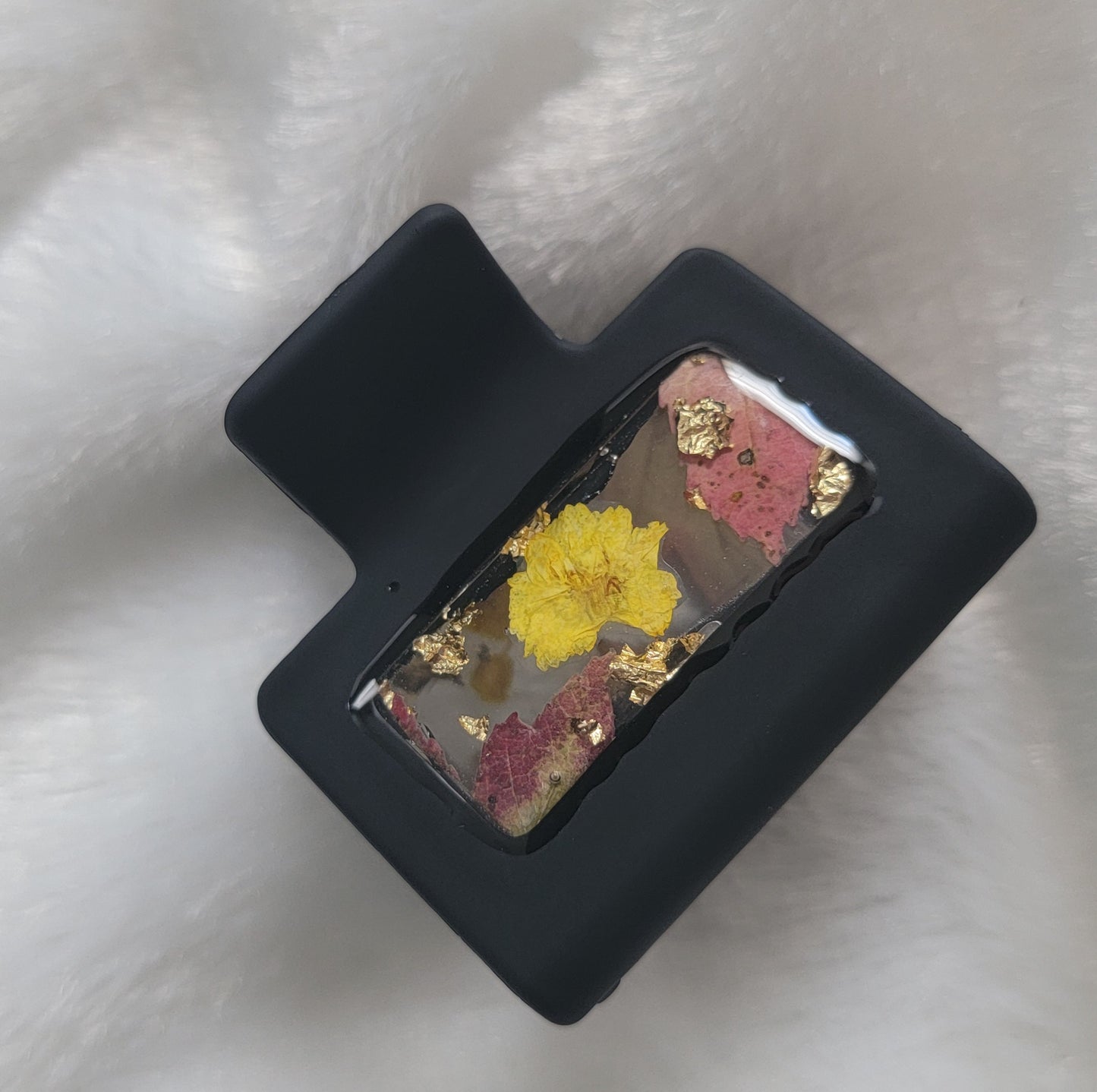 A black 2-inch claw clip with yellow pressed flowers and fall leaves cast in resin within an open space in the hair clip.