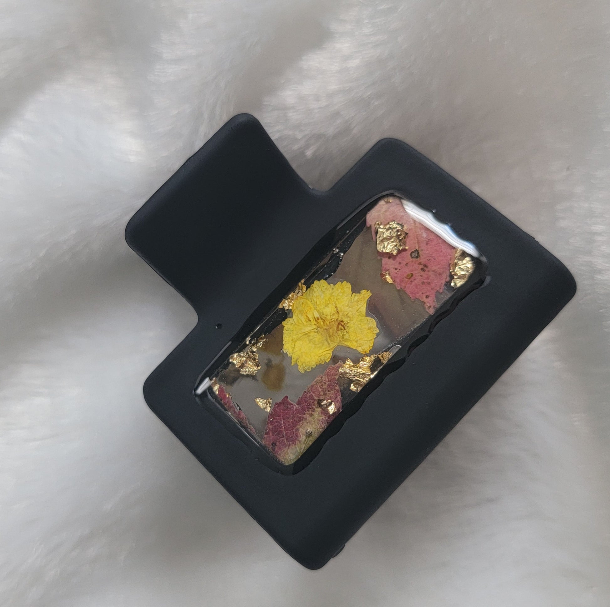 A black 2-inch claw clip with yellow pressed flowers and fall leaves cast in resin within an open space in the hair clip.