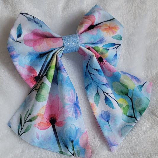 A handmade pet bow with a soft cotton print of watercolor florals in light blues pinks and greens, light blue glitter center sailor bow style with long showy tails.