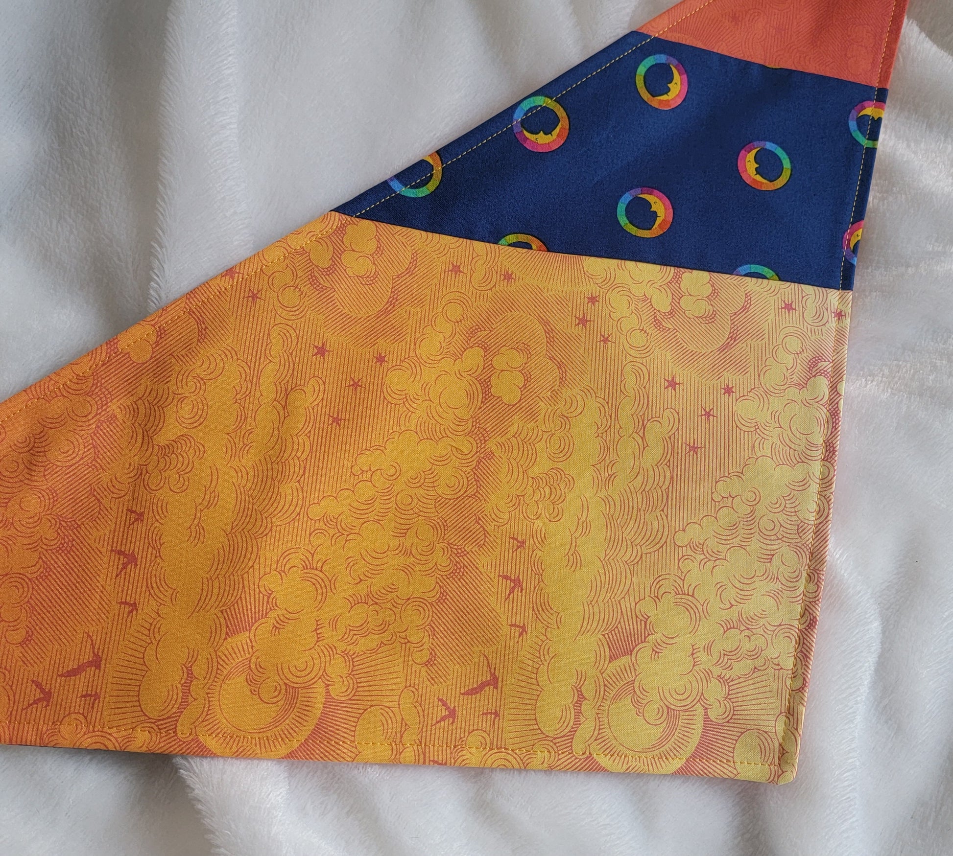 A multicolored pet bandana with stripes of cotton fabric, featuring a soft orange ombre with subtle sky design and contrasting deep blue stripe with rainbow spectrum moons. Laying flat showing side that has more light yellow-orange.