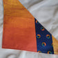 A multicolored pet bandana with stripes of cotton fabric, featuring a soft orange ombre with subtle sky design and contrasting deep blue stripe with rainbow spectrum moons. Laying flat showing side that has more deep orange.