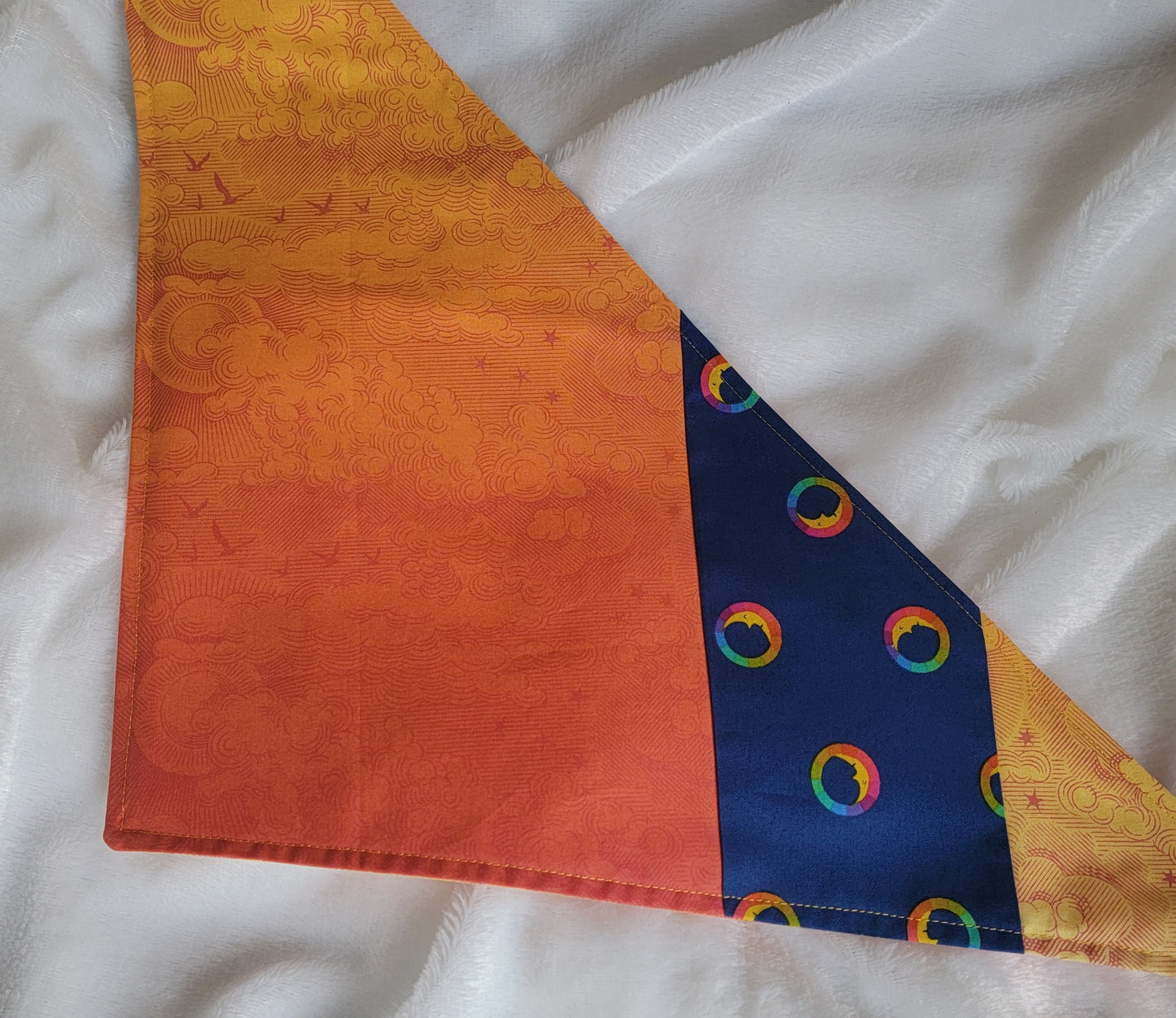 A multicolored pet bandana with stripes of cotton fabric, featuring a soft orange ombre with subtle sky design and contrasting deep blue stripe with rainbow spectrum moons. Laying flat showing side that has more deep orange.