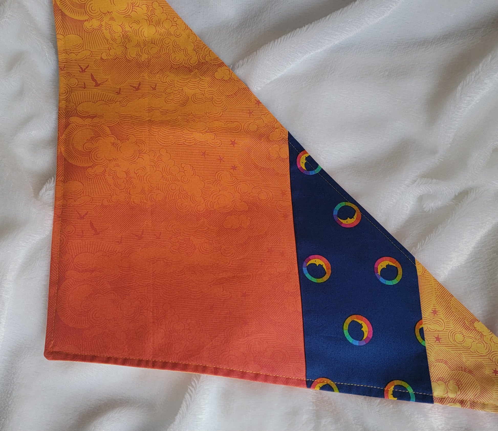 A multicolored pet bandana with stripes of cotton fabric, featuring a soft orange ombre with subtle sky design and contrasting deep blue stripe with rainbow spectrum moons. Laying flat showing side that has more deep orange.