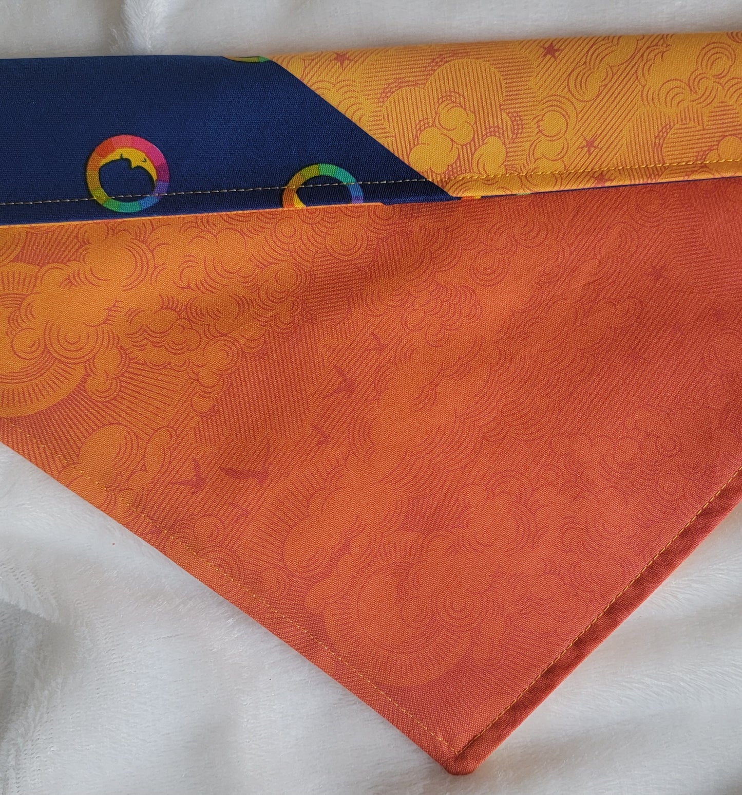 A multicolored pet bandana with stripes of cotton fabric, featuring a soft orange ombre with subtle sky design and contrasting deep blue stripe with rainbow spectrum moons. Showing side that has more deep orange, top edge is folded over to show the reverse side.