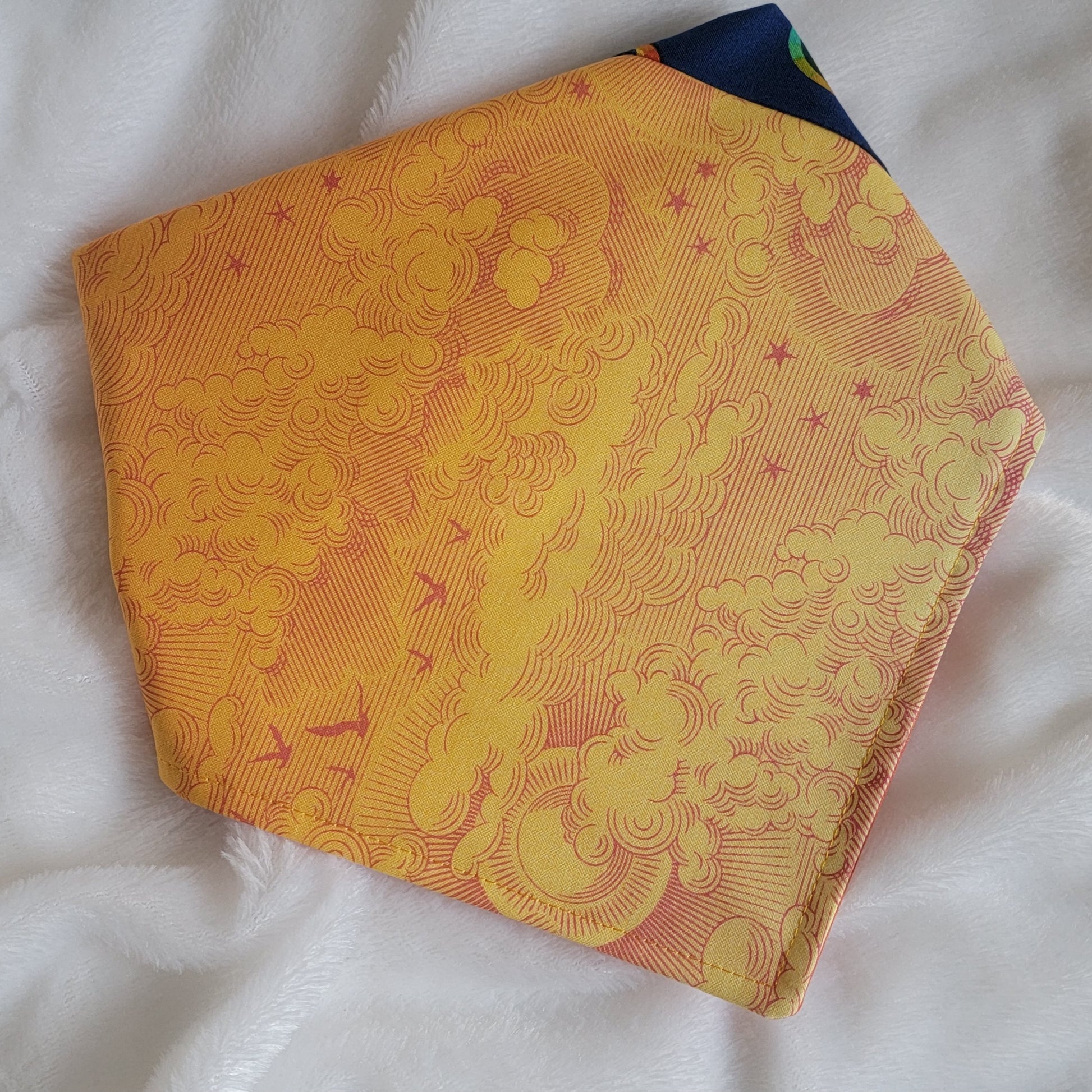 A multicolored pet bandana with stripes of cotton fabric, featuring a soft orange ombre with subtle sky design and contrasting deep blue stripe with rainbow spectrum moons. Corners folded under showing side that has more light yellow-orange.