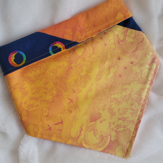A multicolored pet bandana with stripes of cotton fabric, featuring a soft orange ombre with subtle sky design and contrasting deep blue stripe with rainbow spectrum moons. Showing side that has more light yellow-orange, with corners folded under and top edge folded over to show the reverse side.