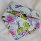 A reversible pet bandana with one side featuring multicolor dragonflies on soft aqua pastel background and the other side vibrant pink and purple waterlilies with dragonflies. Bandana is displayed on white background with corners folded under, showing the side with pink and purple waterlilies with the top edge folded over to show the reverse side as well.