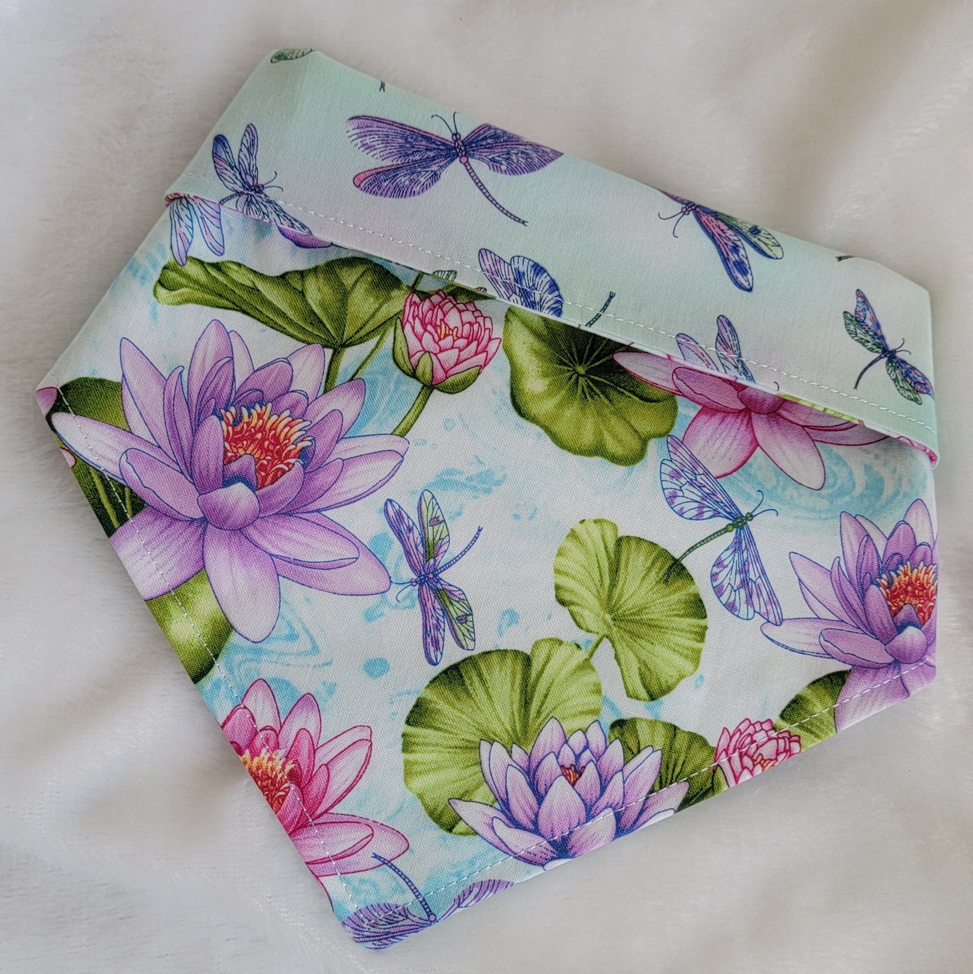 A reversible pet bandana with one side featuring multicolor dragonflies on soft aqua pastel background and the other side vibrant pink and purple waterlilies with dragonflies. Bandana is displayed on white background with corners folded under, showing the side with pink and purple waterlilies with the top edge folded over to show the reverse side as well.