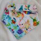 Handmade bandana featuring colorful tropical birds and floral prints patched together in stripes, unique on each side. Displayed with the corners folded under and the top edge folded over to show the reverse side, on a white background.
