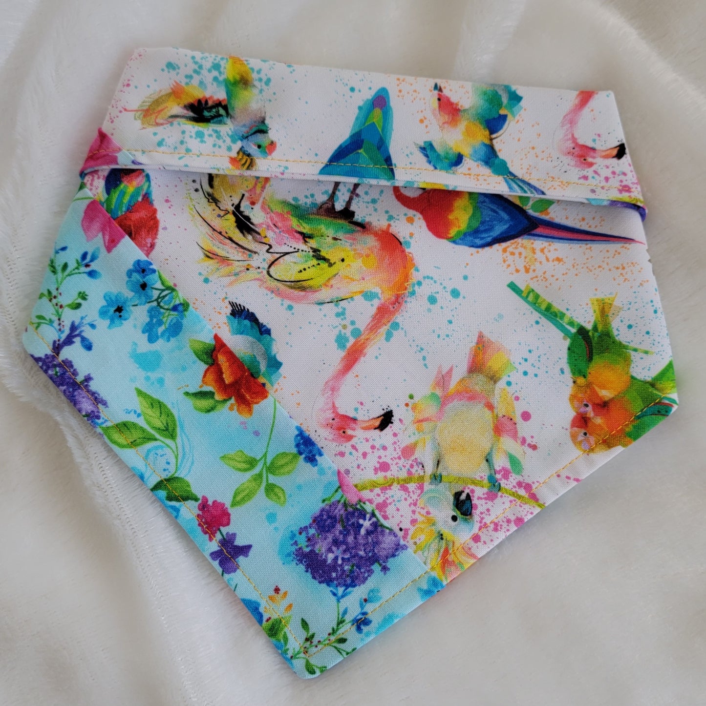 Handmade bandana featuring colorful tropical birds and floral prints patched together in stripes, unique on each side. Displayed with the corners folded under and the top edge folded over to show the reverse side, on a white background.