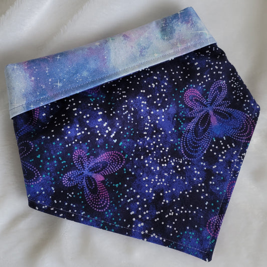 A reversible pet bandana with one side featuring geometric dot patterns that form butterfly shapes in shades of deep purple and the other side soft pastel purple and blue space clouds with silver shimmer. Showing the butterfly side with the corners folded under and top edge folded over to show the reverse side.