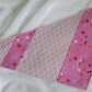 Handmade pet bandana featuring stripes of fabric with lilac scallop pattern and bright pink with cute summer motifs including umbrellas, ice cream, sunglasses, dolphins and more. Laying flat showing side with wider edge stripe of bright pink.