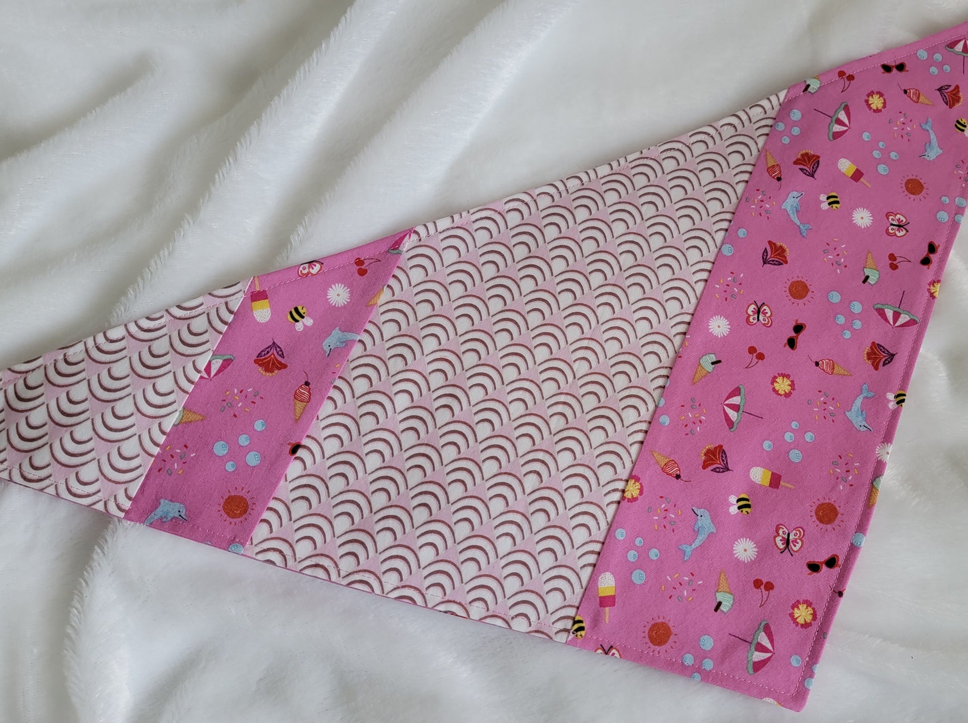 Handmade pet bandana featuring stripes of fabric with lilac scallop pattern and bright pink with cute summer motifs including umbrellas, ice cream, sunglasses, dolphins and more. Laying flat showing side with wider edge stripe of bright pink.