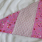 Handmade pet bandana featuring stripes of fabric with lilac scallop pattern and bright pink with cute summer motifs including umbrellas, ice cream, sunglasses, dolphins and more. Laying flat showing side with narrower edge stripe of bright pink.