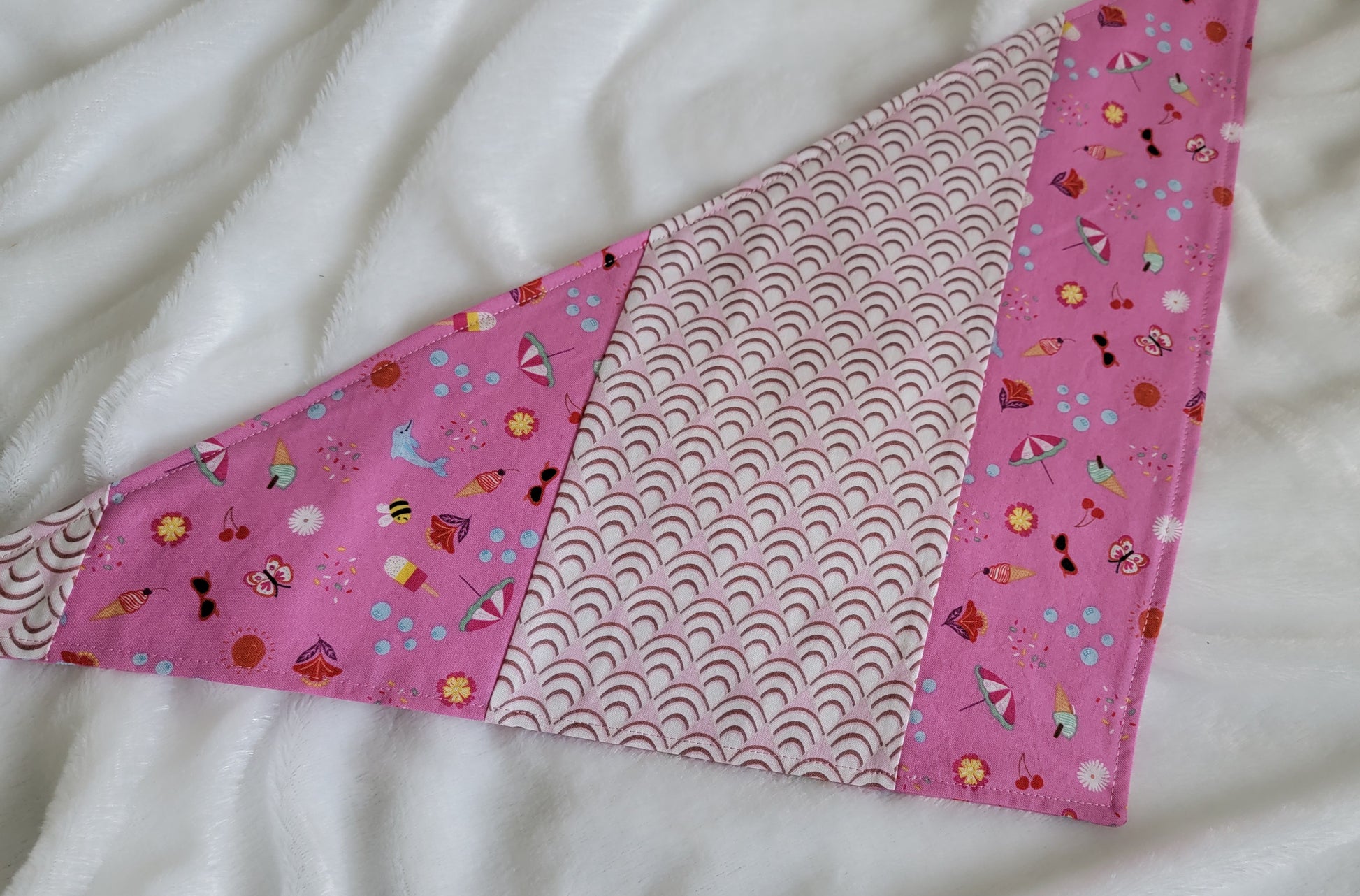 Handmade pet bandana featuring stripes of fabric with lilac scallop pattern and bright pink with cute summer motifs including umbrellas, ice cream, sunglasses, dolphins and more. Laying flat showing side with narrower edge stripe of bright pink.