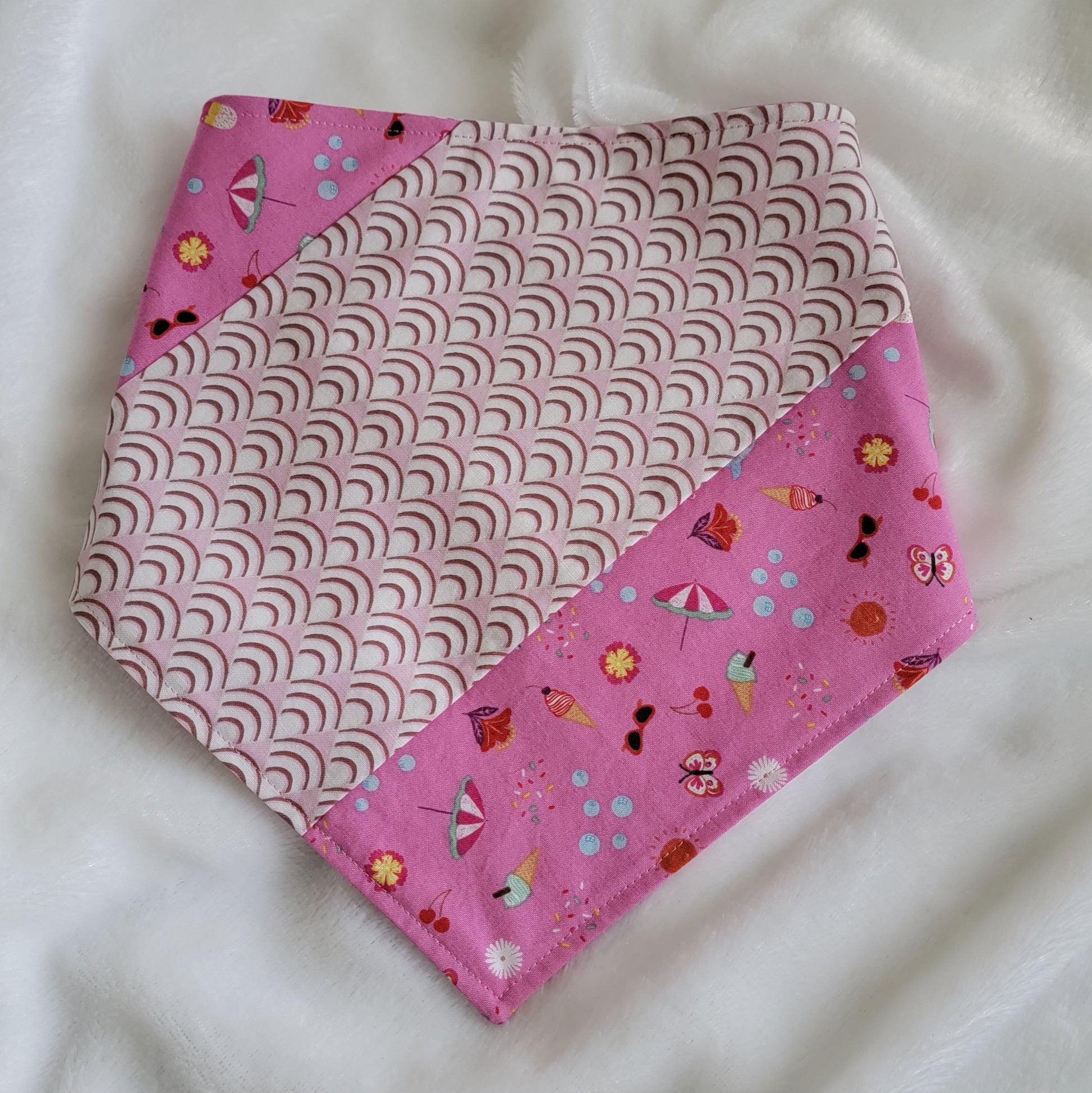 Handmade pet bandana featuring stripes of fabric with lilac scallop pattern and bright pink with cute summer motifs including umbrellas, ice cream, sunglasses, dolphins and more. Showing side with narrower edge stripe of bright pink, corners are folded under.