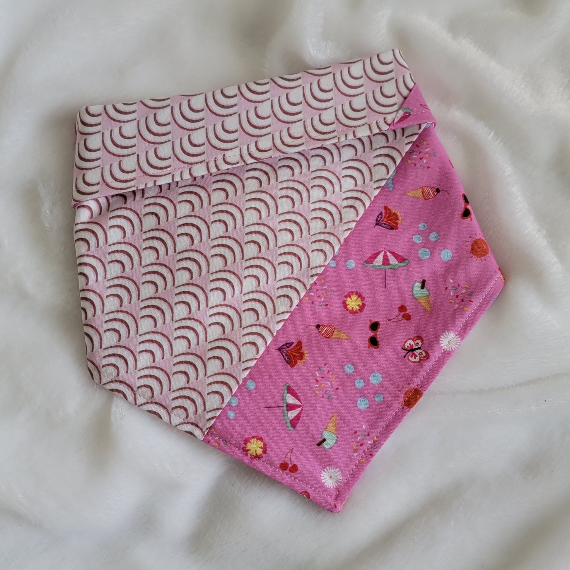 Handmade pet bandana featuring stripes of fabric with lilac scallop pattern and bright pink with cute summer motifs including umbrellas, ice cream, sunglasses, dolphins and more. Showing side with narrower edge stripe of bright pink, corners are folded under and top edge is folded over to show the reverse side.