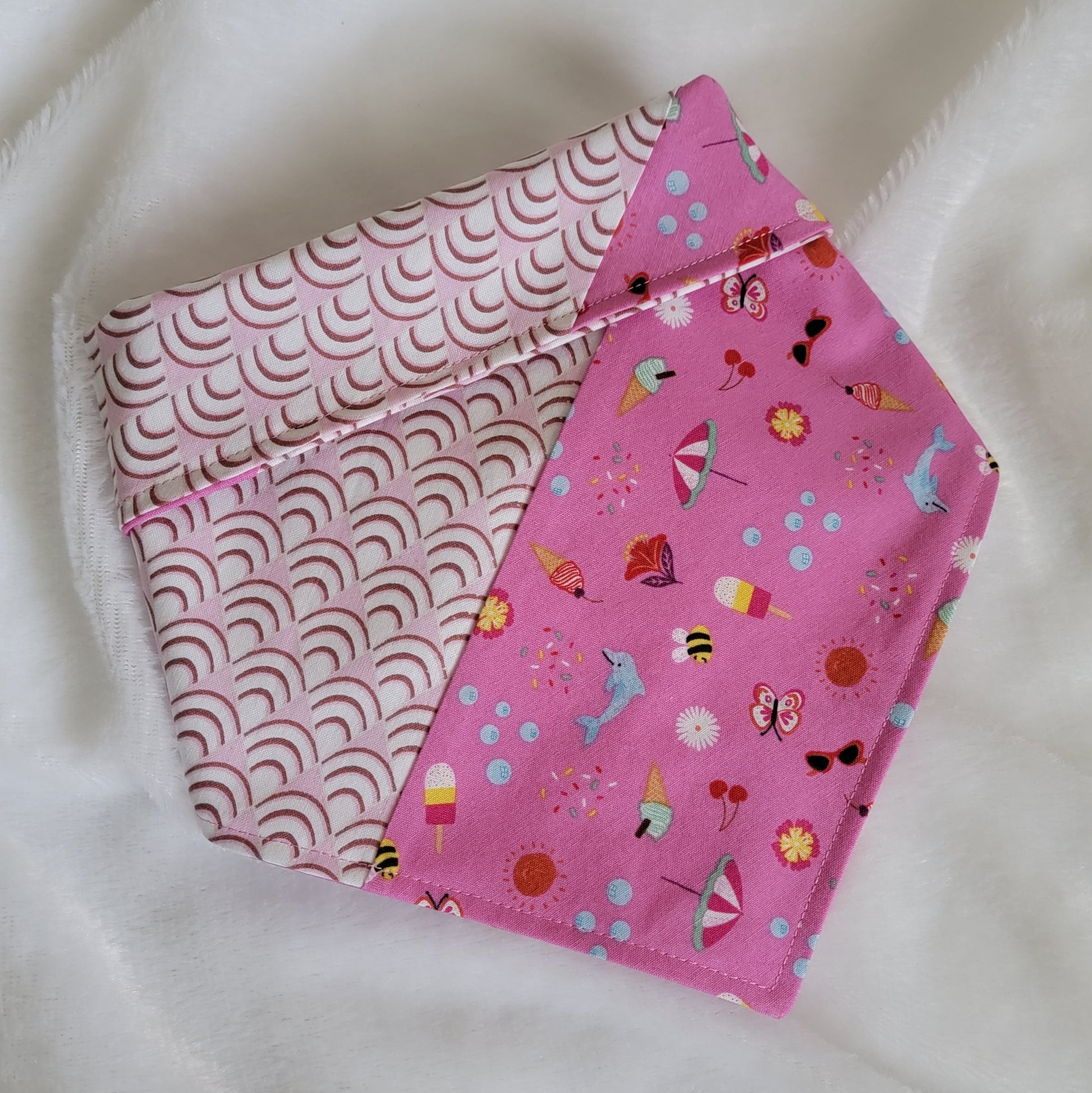 Handmade pet bandana featuring stripes of fabric with lilac scallop pattern and bright pink with cute summer motifs including umbrellas, ice cream, sunglasses, dolphins and more. Showing side with wider edge stripe of bright pink, corners are folded under and top edge is folded over to show the reverse side.