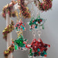 Two pairs of drop earrings, green and red and Christmas glitters in star shaped resin pieces with gold colored and silver colored metal findings. Earrings are displayed on a white earring hanger with red and gold garland wrapped around it.