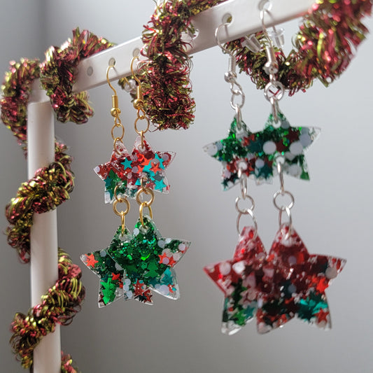 Two pairs of drop earrings, green and red and Christmas glitters in star shaped resin pieces with gold colored and silver colored metal findings. Earrings are displayed on a white earring hanger with red and gold garland wrapped around it.