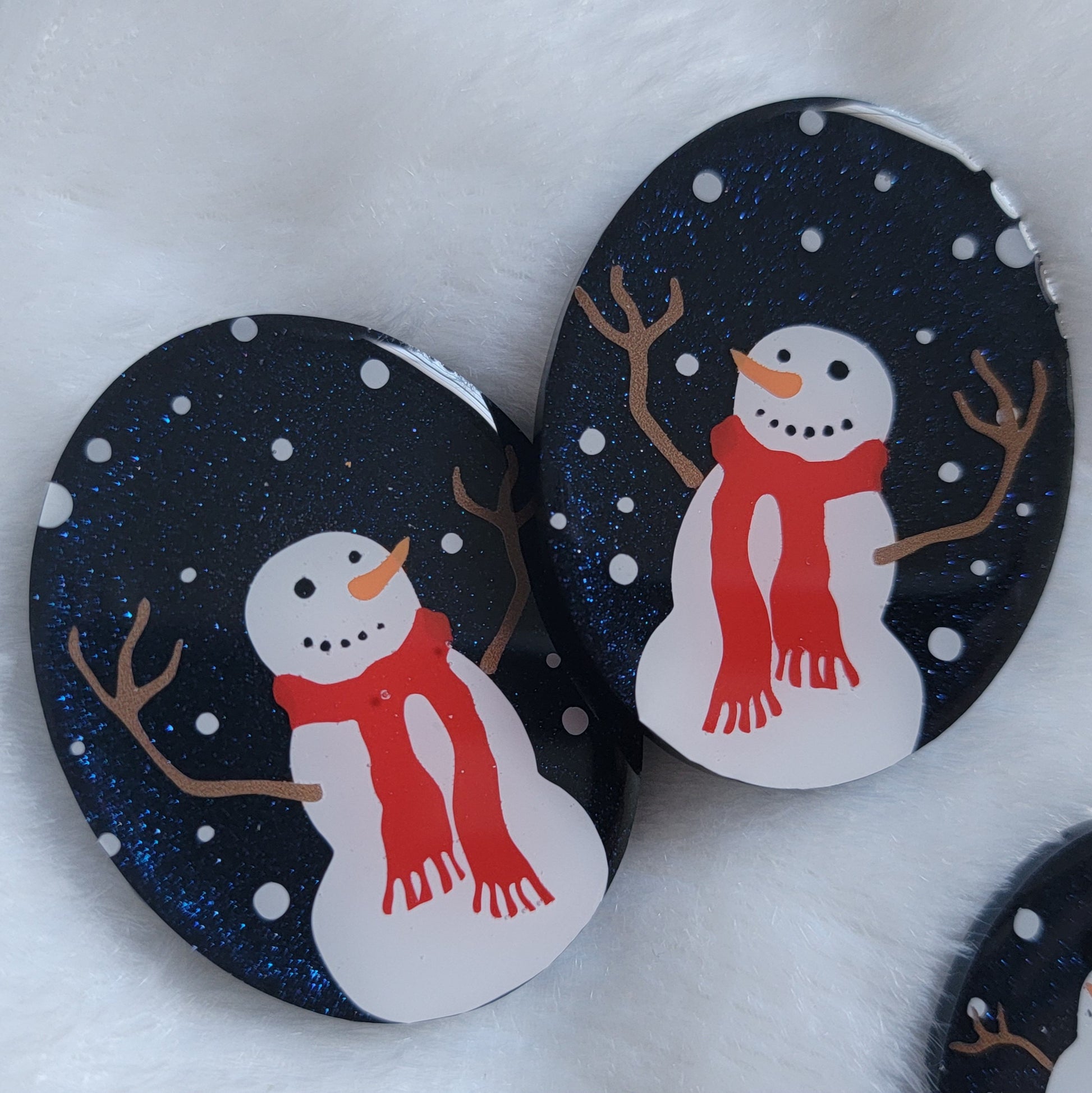 Two dark blue sparkly oval-shaped epoxy resin pieces with snowman designs, displayed on a white background