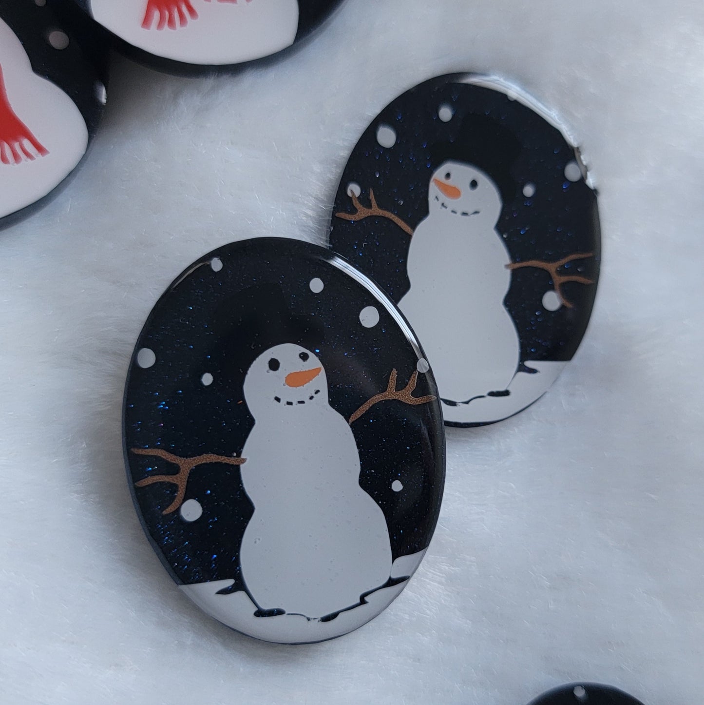 Two dark blue sparkly oval-shaped epoxy resin pieces with snowman designs, displayed on a white background