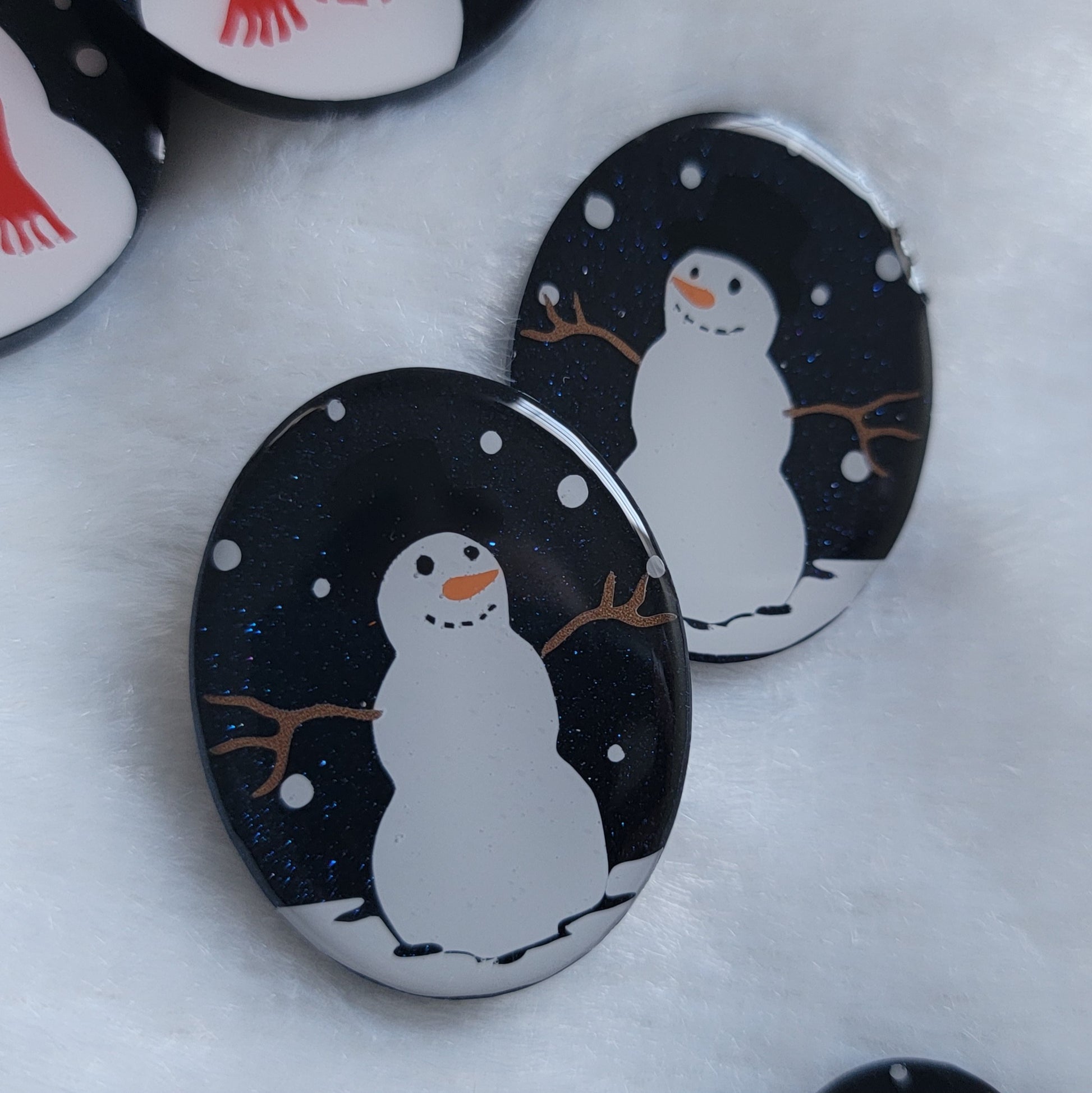 Two dark blue sparkly oval-shaped epoxy resin pieces with snowman designs, displayed on a white background