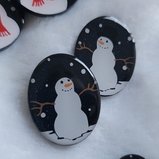 Two dark blue sparkly oval-shaped epoxy resin pieces with snowman designs, displayed on a white background