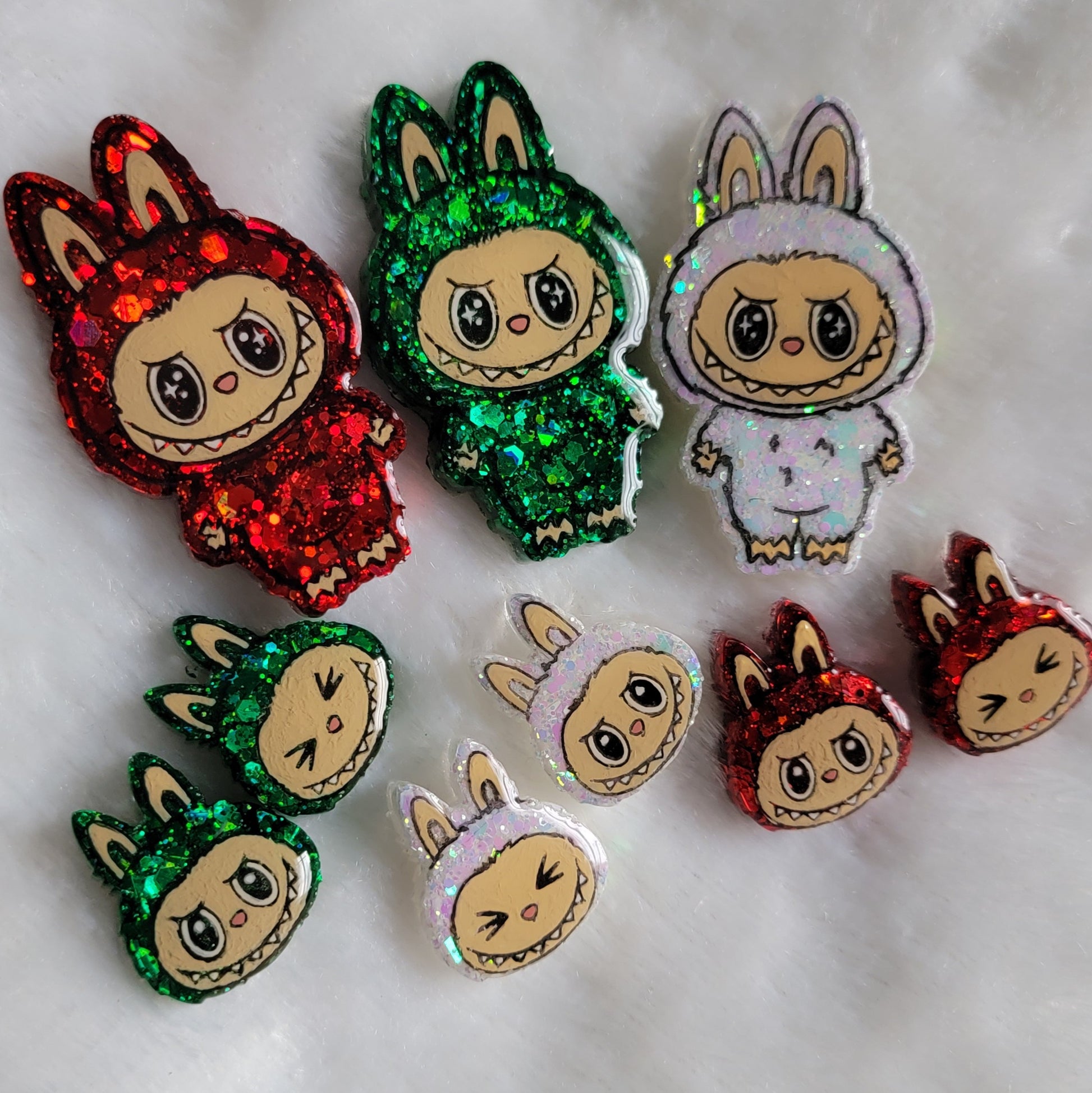 Labubu inspired epoxy resin pet tags, small pieces with just the monster face and larger pieces with the full monster character. They're made with red, green, and iridescent white glitter and acrylic painted details, and displayed on a soft white background.