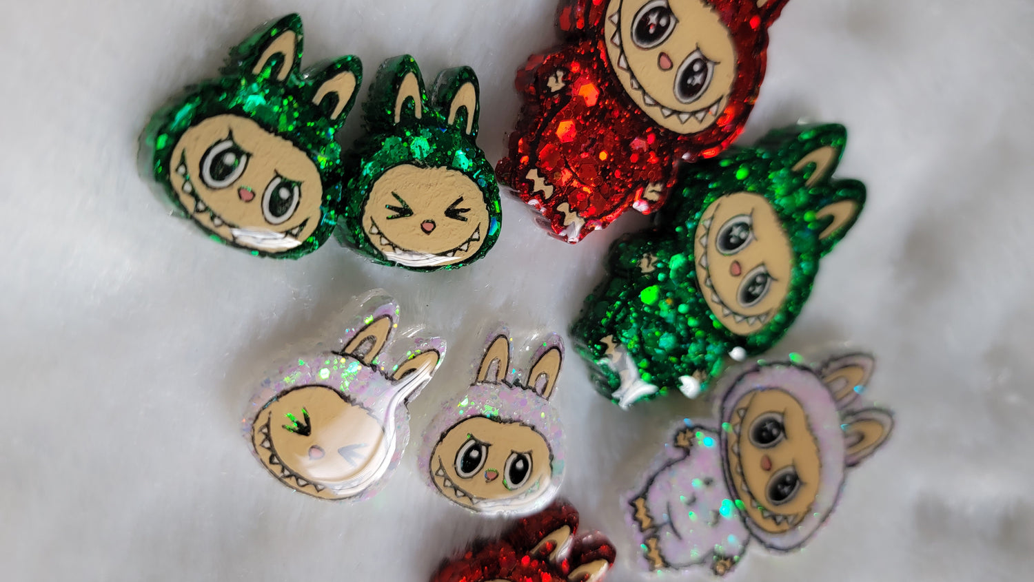 Labubu inspired epoxy resin pet tags, small pieces with just the monster face and larger pieces with the full monster character. They're made with red, green, and iridescent white glitter and acrylic painted details, and displayed on a soft white background.
